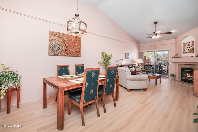 dining area featuring ceiling fan with notable chandelier, a lit fireplace, high vaulted ceiling, and light wood-style flooring