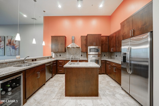 kitchen featuring appliances with stainless steel finishes, wall chimney exhaust hood, beverage cooler, a center island with sink, and hanging light fixtures