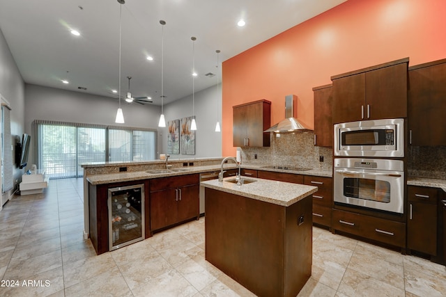 kitchen with wall chimney exhaust hood, stainless steel appliances, wine cooler, an island with sink, and decorative light fixtures