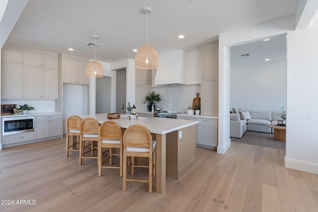 kitchen with pendant lighting, light hardwood / wood-style floors, custom range hood, a center island with sink, and built in microwave