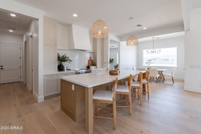 kitchen with tasteful backsplash, custom range hood, a center island with sink, decorative light fixtures, and light wood-type flooring