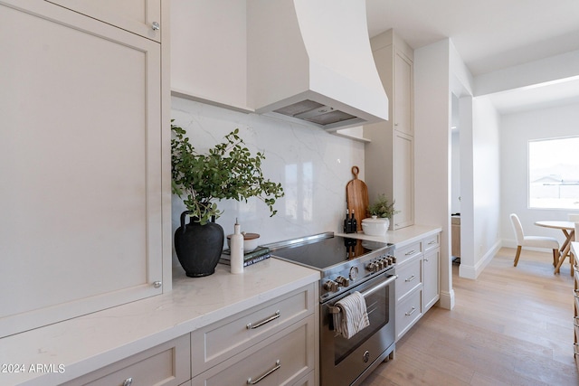 kitchen with custom exhaust hood, light stone counters, high end range, light hardwood / wood-style floors, and decorative backsplash