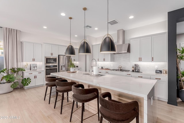 kitchen featuring sink, stainless steel appliances, an island with sink, white cabinets, and wall chimney exhaust hood