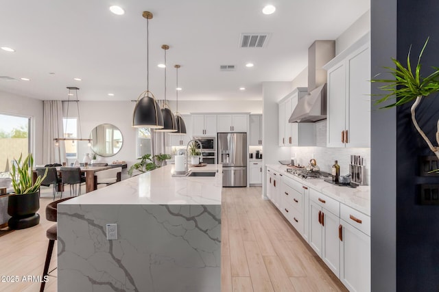 kitchen featuring pendant lighting, wall chimney range hood, white cabinetry, stainless steel appliances, and a large island with sink