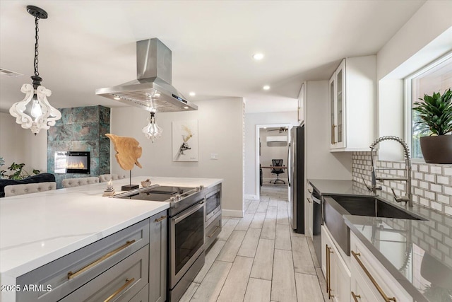 kitchen with gray cabinets, island exhaust hood, appliances with stainless steel finishes, a sink, and glass insert cabinets