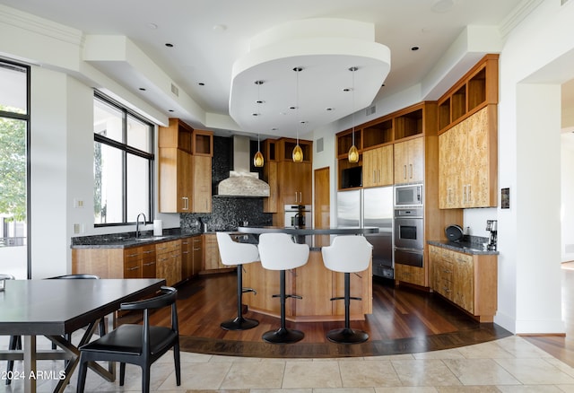 kitchen featuring tasteful backsplash, wall chimney range hood, built in appliances, a kitchen island, and hanging light fixtures