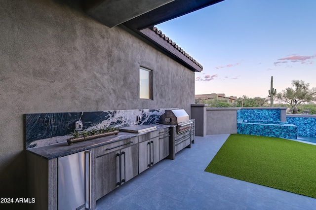 patio terrace at dusk with a grill, a lawn, and exterior kitchen