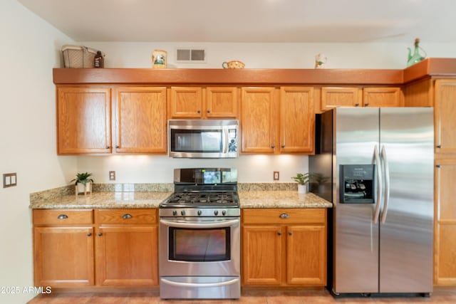kitchen featuring appliances with stainless steel finishes, brown cabinetry, light stone counters, and visible vents