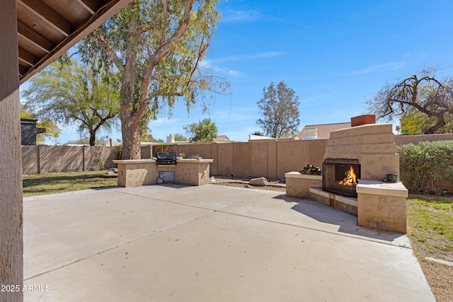 view of patio featuring a fenced backyard, a warm lit fireplace, and an outdoor kitchen