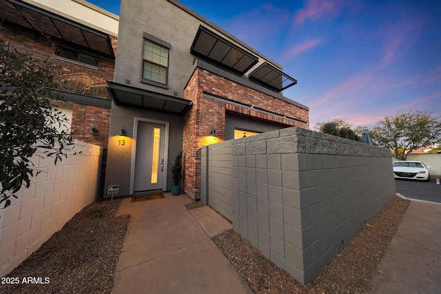 exterior entry at dusk with brick siding, fence, and stucco siding