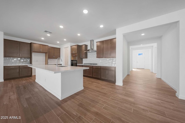 kitchen featuring sink, a kitchen island with sink, wall chimney exhaust hood, oven, and stainless steel gas stovetop