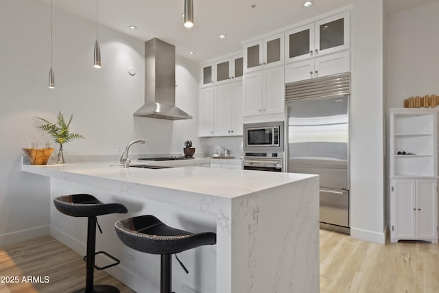 kitchen featuring white cabinetry, hanging light fixtures, range hood, built in appliances, and light stone countertops