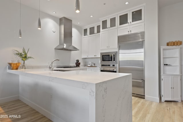 kitchen with hanging light fixtures, range hood, built in appliances, light stone countertops, and white cabinets