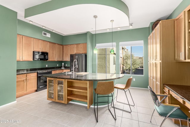 kitchen featuring light tile patterned flooring, decorative light fixtures, dark stone countertops, a kitchen island with sink, and black appliances