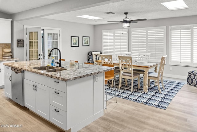 kitchen with stainless steel dishwasher, a ceiling fan, an island with sink, light wood-type flooring, and a sink