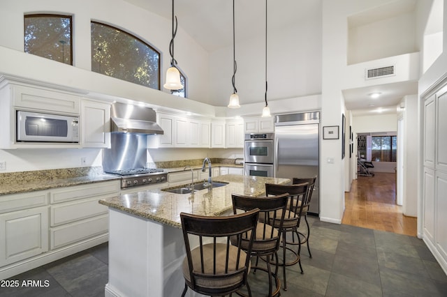 kitchen with light stone counters, built in appliances, sink, and wall chimney range hood