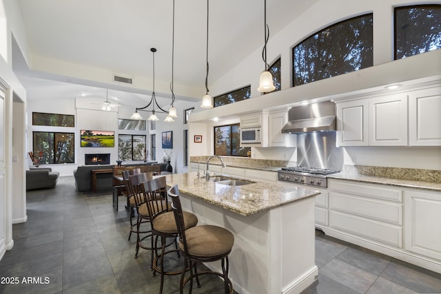 kitchen with pendant lighting, white microwave, sink, a kitchen island with sink, and wall chimney range hood