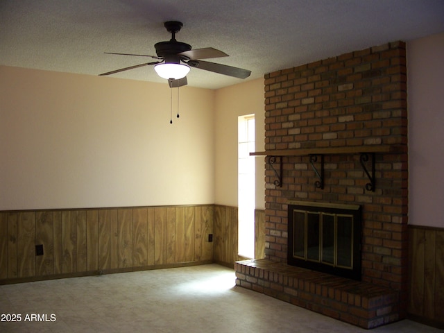 unfurnished living room with wood walls, wainscoting, a fireplace, a textured ceiling, and a ceiling fan