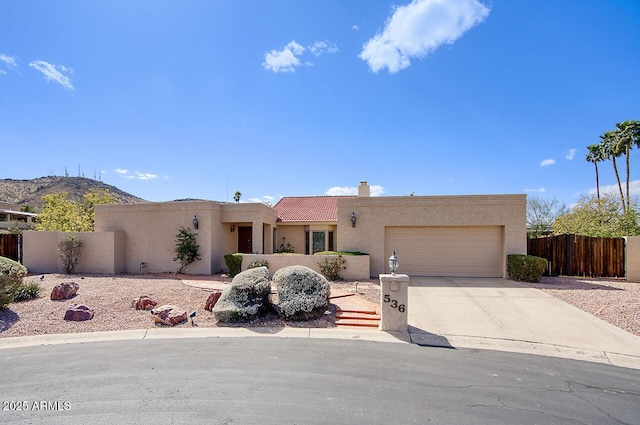 pueblo-style house featuring a garage, concrete driveway, a chimney, fence, and stucco siding