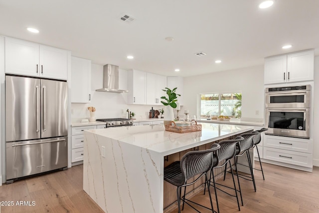 kitchen featuring visible vents, a kitchen island, stainless steel appliances, wall chimney exhaust hood, and light wood finished floors