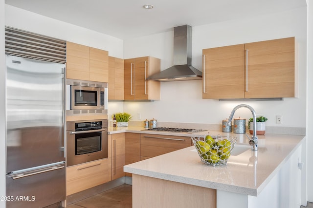 kitchen with built in appliances, wall chimney exhaust hood, a peninsula, light stone counters, and light brown cabinets