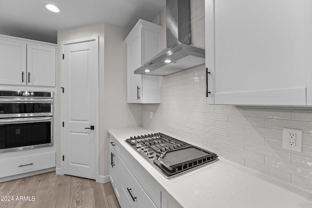 kitchen with white cabinets, tasteful backsplash, wall chimney range hood, appliances with stainless steel finishes, and light wood-type flooring