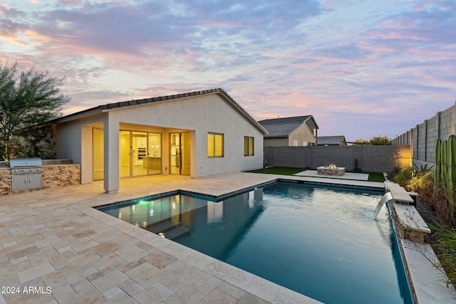 pool at dusk with a patio, a fire pit, exterior kitchen, a grill, and pool water feature