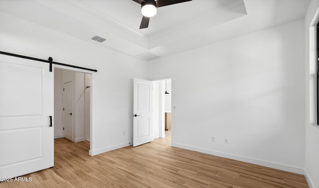 unfurnished bedroom with a tray ceiling, a barn door, light hardwood / wood-style floors, and ceiling fan