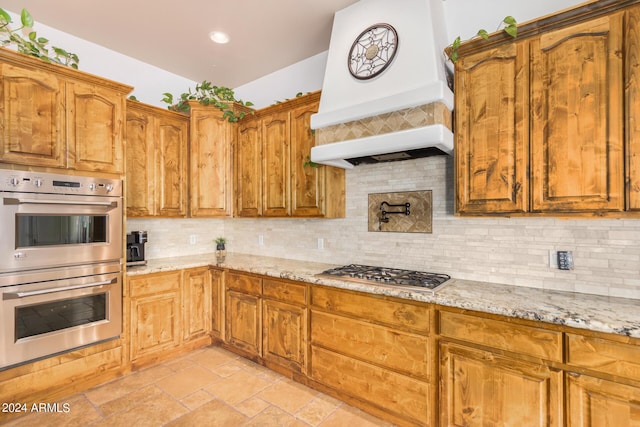kitchen with backsplash, custom exhaust hood, stainless steel appliances, and light stone countertops