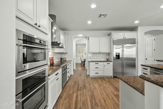 kitchen featuring arched walkways, wood finished floors, stainless steel appliances, white cabinetry, and exhaust hood