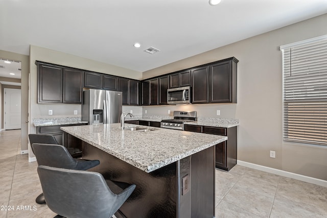 kitchen with an island with sink, appliances with stainless steel finishes, light stone countertops, dark brown cabinetry, and sink