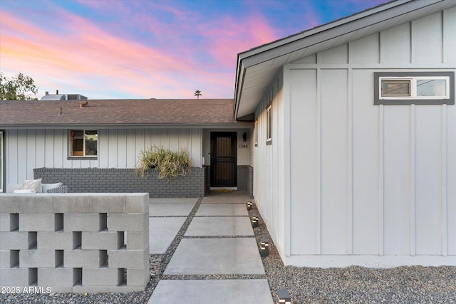 exterior entry at dusk featuring board and batten siding, brick siding, and a shingled roof