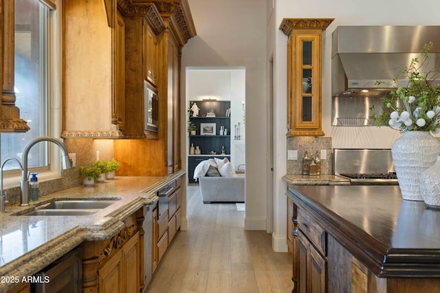 kitchen featuring a sink, light wood-style floors, wall chimney range hood, backsplash, and brown cabinetry