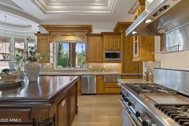 kitchen with under cabinet range hood, stainless steel appliances, a sink, brown cabinetry, and a raised ceiling