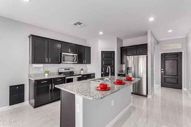 kitchen featuring visible vents, a center island with sink, a sink, stainless steel appliances, and light stone countertops