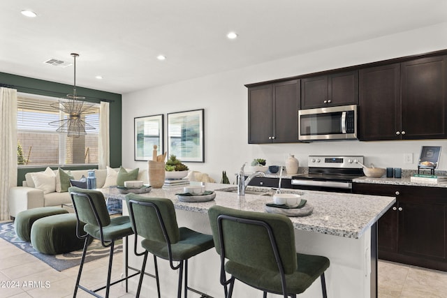 kitchen with sink, appliances with stainless steel finishes, a kitchen island with sink, light stone countertops, and decorative light fixtures