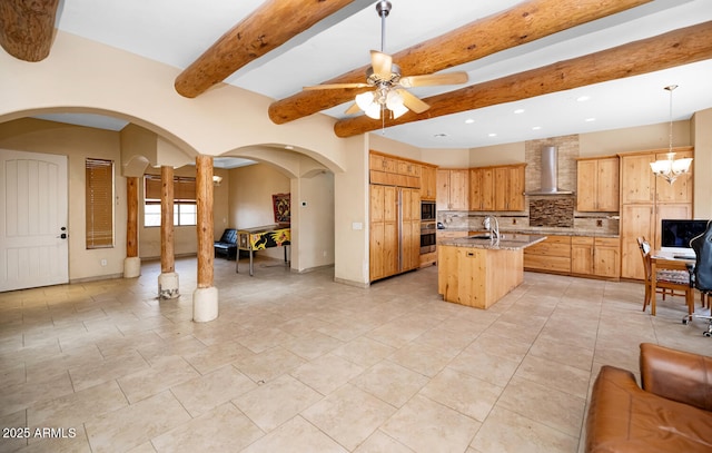 kitchen featuring light stone counters, a center island with sink, decorative backsplash, wall chimney range hood, and ceiling fan with notable chandelier