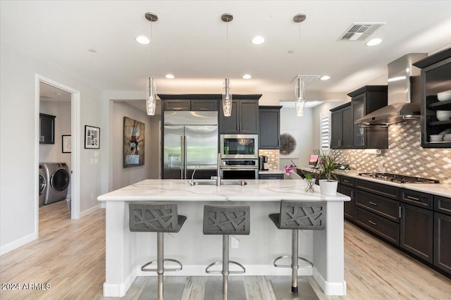 kitchen with wall chimney range hood, built in appliances, pendant lighting, light hardwood / wood-style floors, and a kitchen island with sink