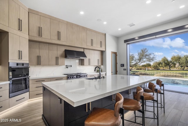 kitchen featuring sink, light brown cabinets, a kitchen breakfast bar, range hood, and a kitchen island with sink