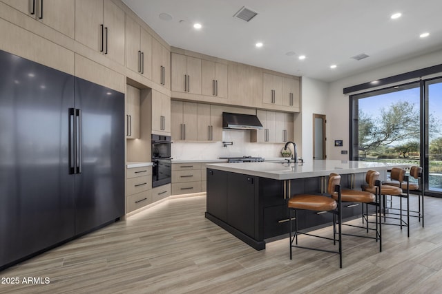 kitchen featuring a center island with sink, tasteful backsplash, black appliances, a kitchen bar, and wall chimney exhaust hood