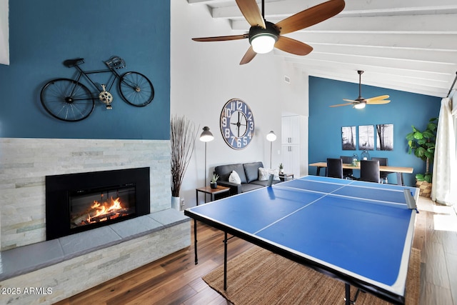 rec room featuring vaulted ceiling with beams, a ceiling fan, wood finished floors, and a stone fireplace