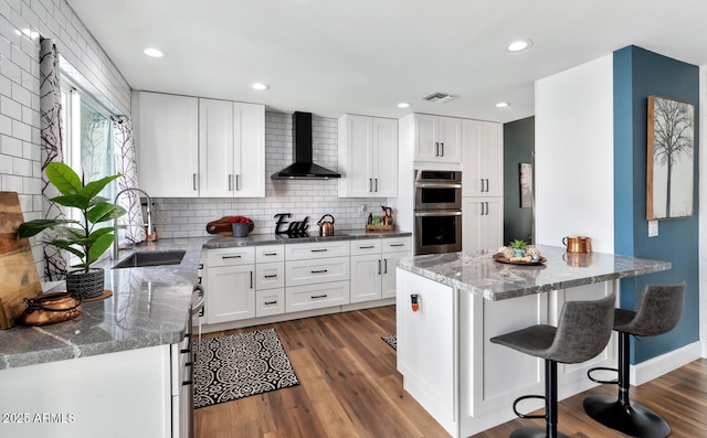 kitchen featuring dark wood finished floors, visible vents, stainless steel double oven, a sink, and wall chimney range hood