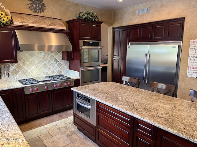 kitchen with light stone countertops, appliances with stainless steel finishes, wall chimney range hood, and backsplash