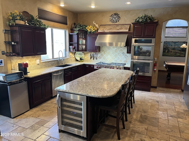 kitchen featuring wall chimney range hood, sink, stainless steel appliances, a center island, and beverage cooler