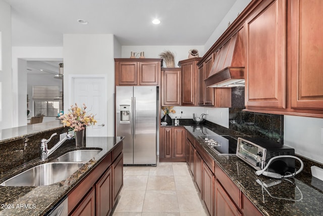 kitchen with sink, premium range hood, dark stone countertops, stainless steel fridge, and light tile patterned floors