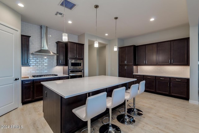 kitchen featuring wall chimney range hood, a center island, stainless steel appliances, light wood finished floors, and light countertops