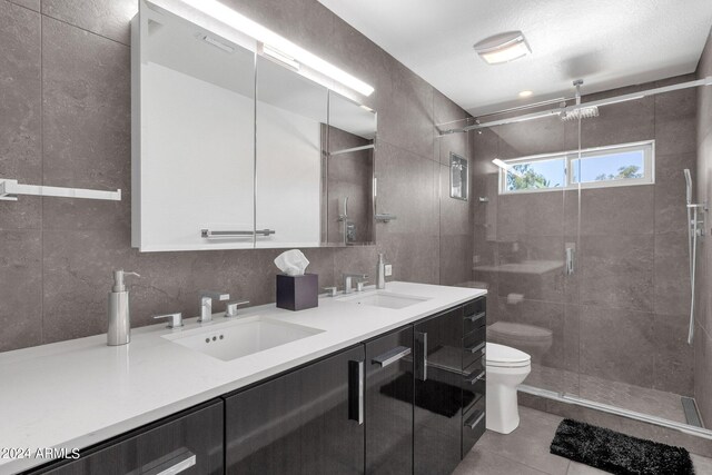 bathroom with double sink vanity, tile walls, toilet, a tile shower, and tile flooring