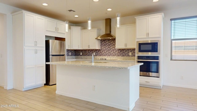 kitchen featuring decorative backsplash, a center island with sink, wall chimney range hood, light wood finished floors, and stainless steel appliances