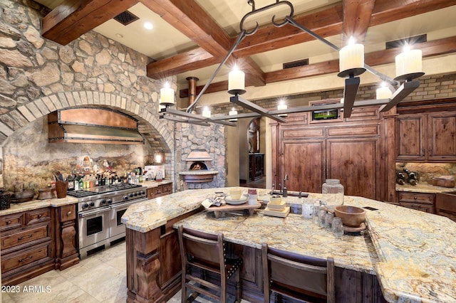 kitchen featuring an island with sink, beam ceiling, light stone counters, range with two ovens, and a breakfast bar area