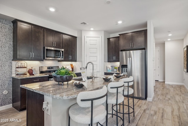 kitchen with sink, a kitchen island with sink, stainless steel appliances, light stone counters, and dark brown cabinetry